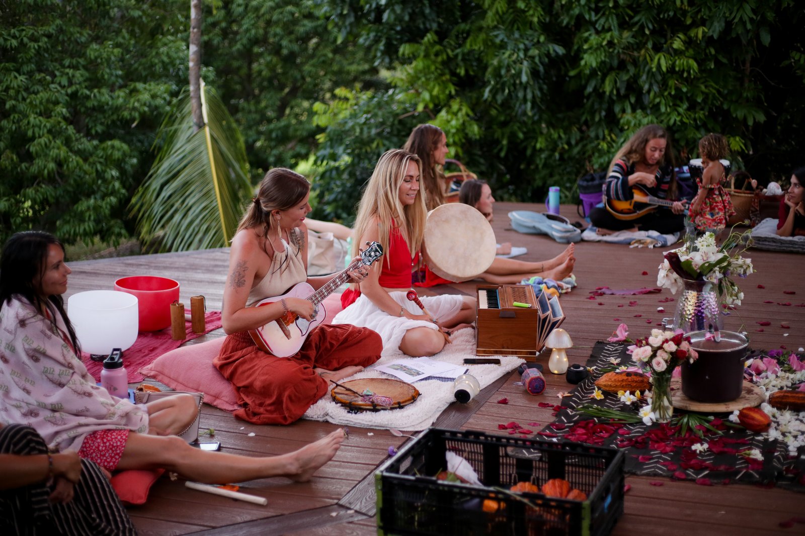 A group gathered for a sound healing ceremony with ukulele, frame drum, harmonium, and singing bowls on a deck surrounded by jungle
